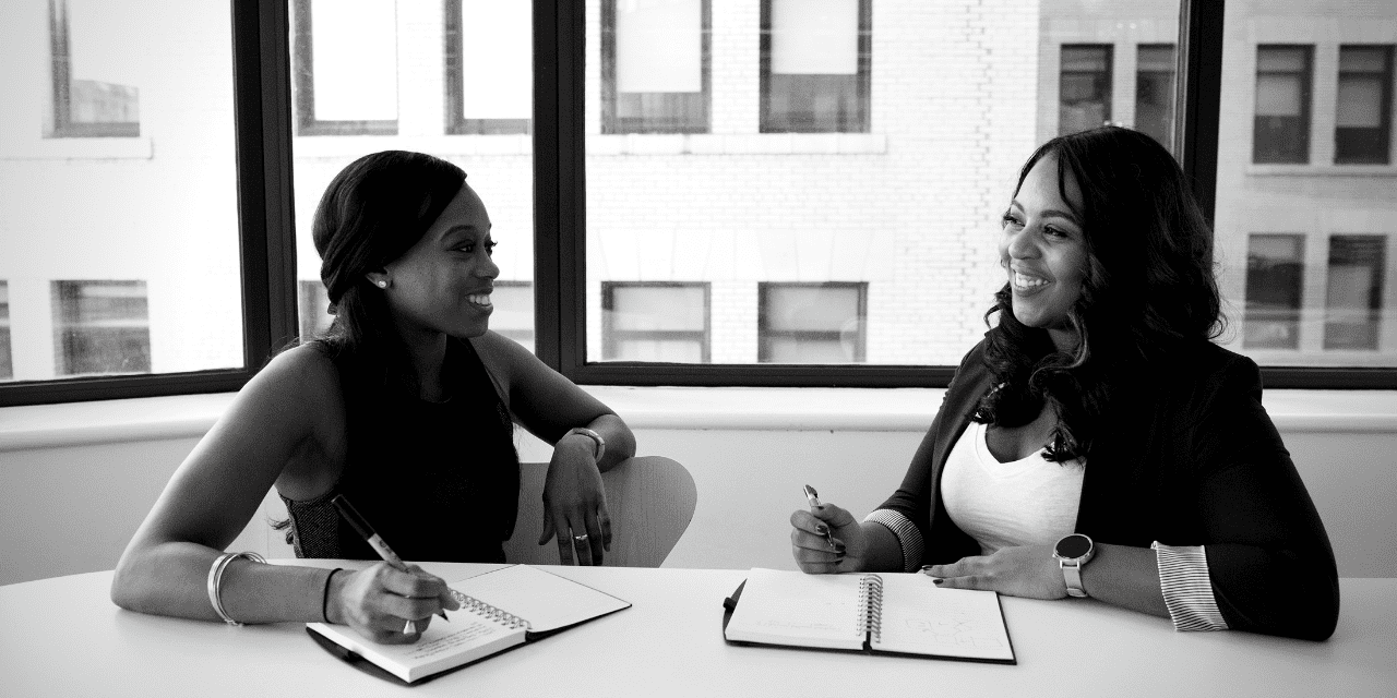 Two women engaged in discussion at a table, with a notebook open between them, sharing ideas and collaborating.