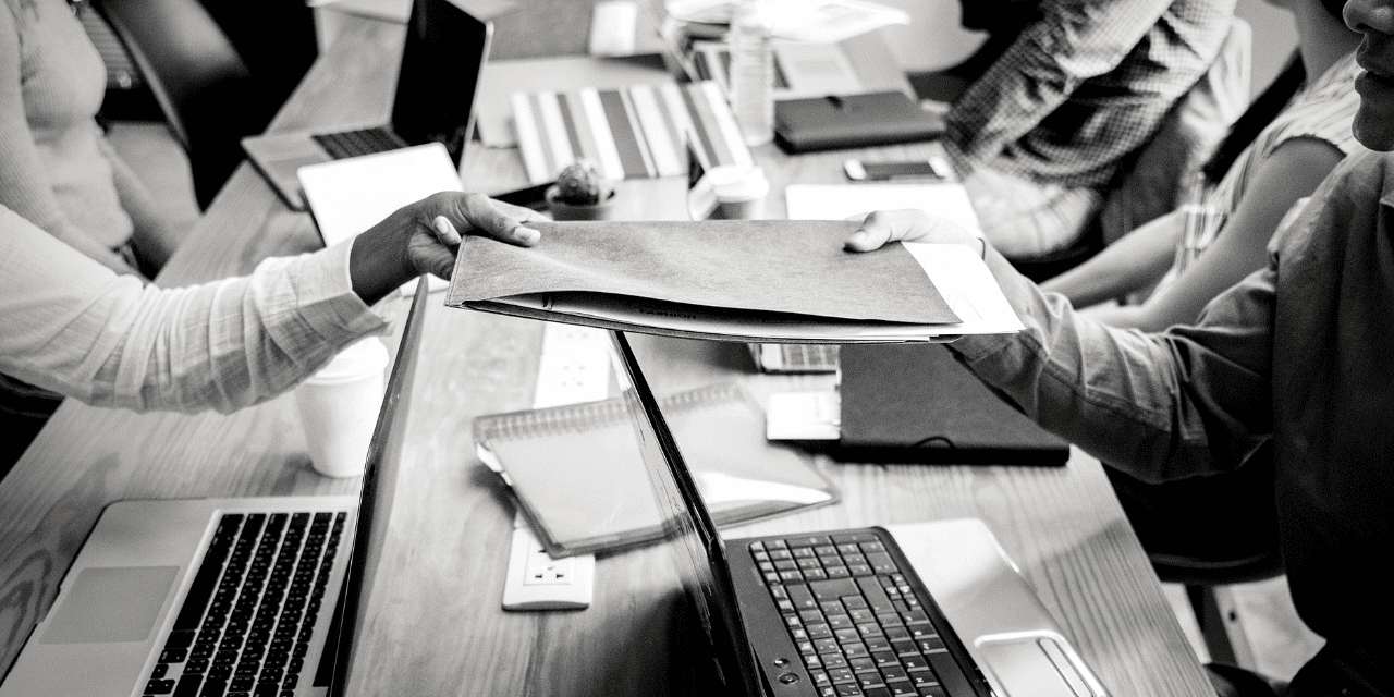 A monochrome image of a person handing over a folder across a busy office desk with laptops and papers.