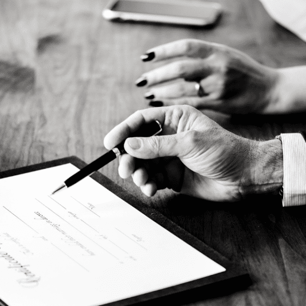 Person signing a document with another observing; focus on hands, black and white image.